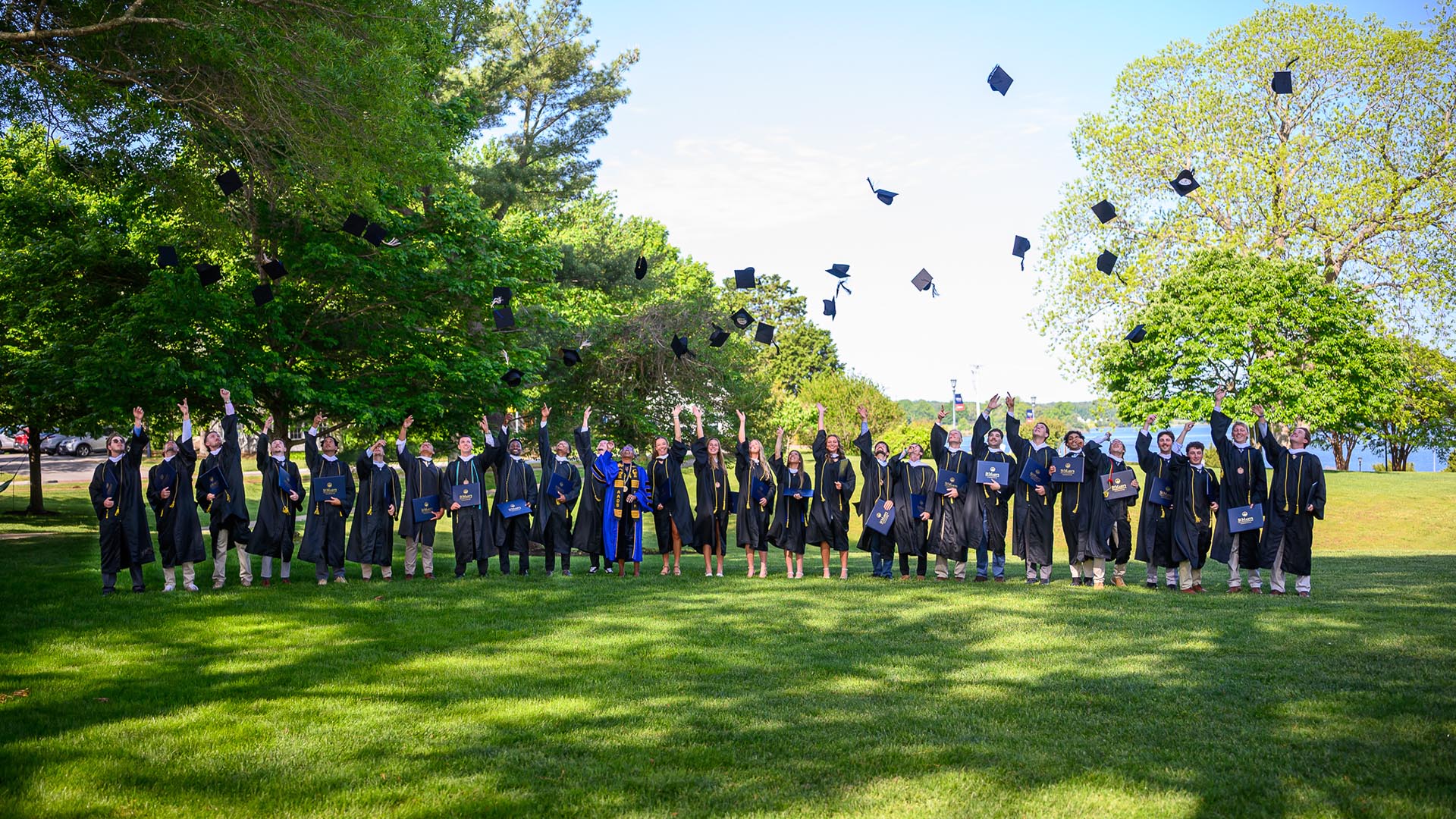 A group of graduates in caps and gowns stand on a lawn, tossing their caps into the air in celebration, surrounded by green trees under a clear sky.