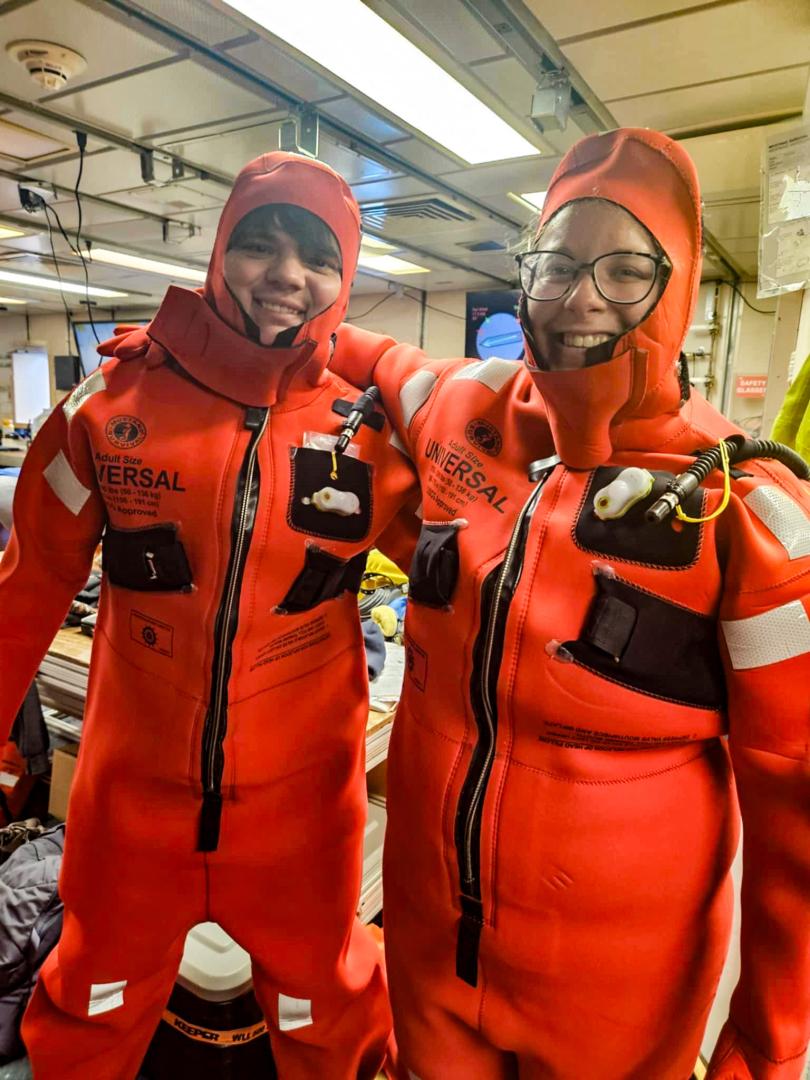 Two people wearing orange immersion survival suits smile inside a room filled with equipment and supplies.