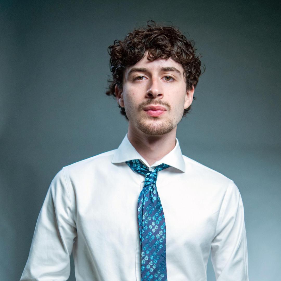 A young man with curly hair wearing a white shirt and blue patterned tie stands against a plain gray background, looking directly at the camera.
