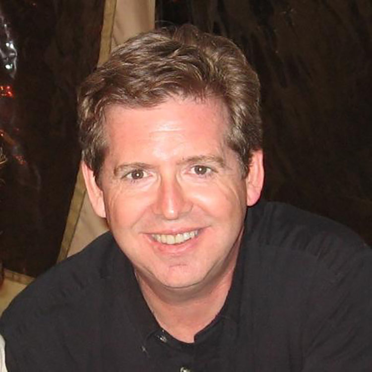 A man with short brown hair and a black shirt smiles at the camera in an indoor setting.