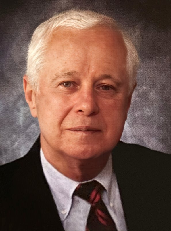An older man with short white hair, wearing a black suit, white shirt, and red-and-black striped tie, poses against a textured gray background.