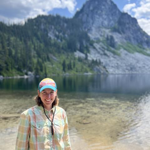 A person wearing a plaid shirt and colorful cap stands near a clear mountain lake with a forested, rocky peak in the background.