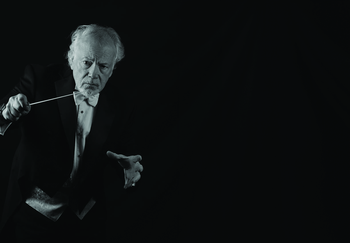 An older man in formal attire holds a conductor’s baton and poses against a dark background, appearing to conduct music.