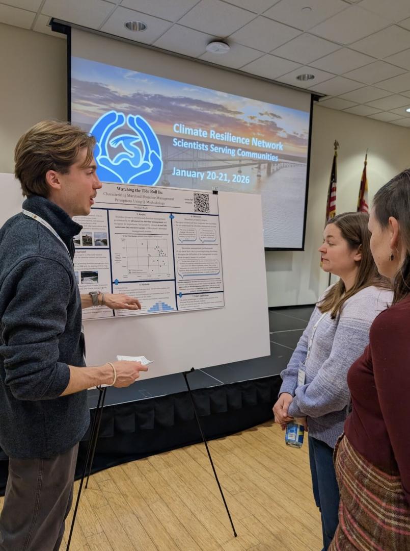 A student presents a research poster to two women with a display screen and American flags visible in the background.
