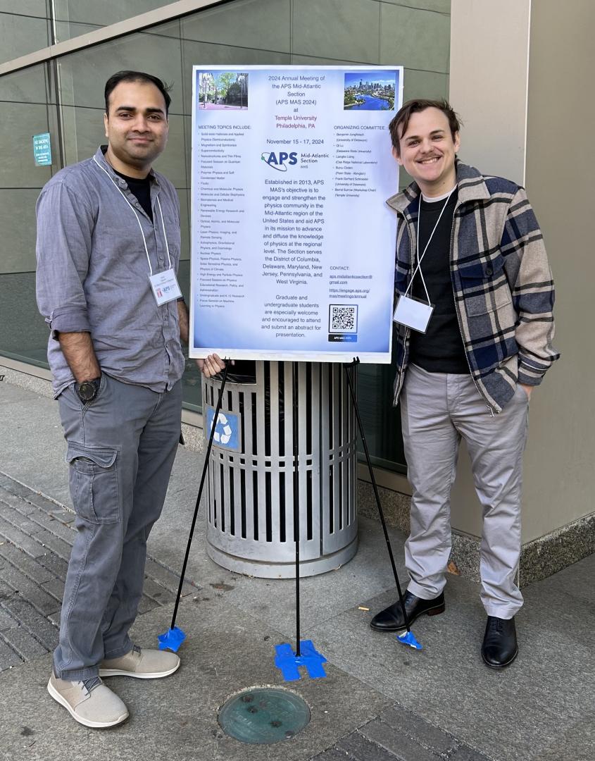 Anish Agashe (left) and Alex Stornelli (right) stand on either side of a conference poster displayed on a stand outdoors, both smiling at the camera.