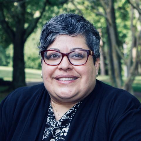 A person with short gray hair and glasses smiles outdoors, wearing a black and white patterned top and a black jacket. Trees and greenery are in the background.