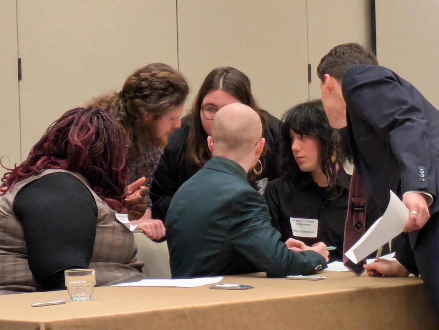 SMCM ethics bowl team  seated and standing around a table, closely discussing ethics in a conference or meeting room.