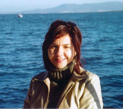 A woman with dark hair and a beige jacket stands in front of a large body of water with distant mountains in the background.