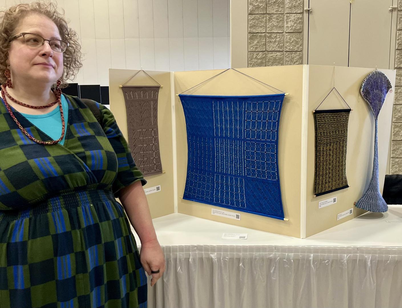 A woman stands next to a display featuring four geometric textile artworks and a tall, curved sculptural piece on a table.