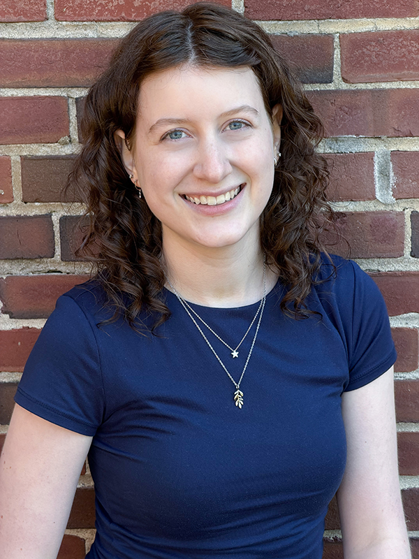 A woman with curly brown hair wearing a navy blue shirt and layered necklaces stands smiling in front of a brick wall.