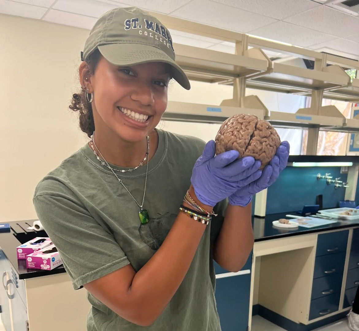 A woman wearing gloves and a cap holds a brain specimen in a laboratory setting, smiling at the camera.