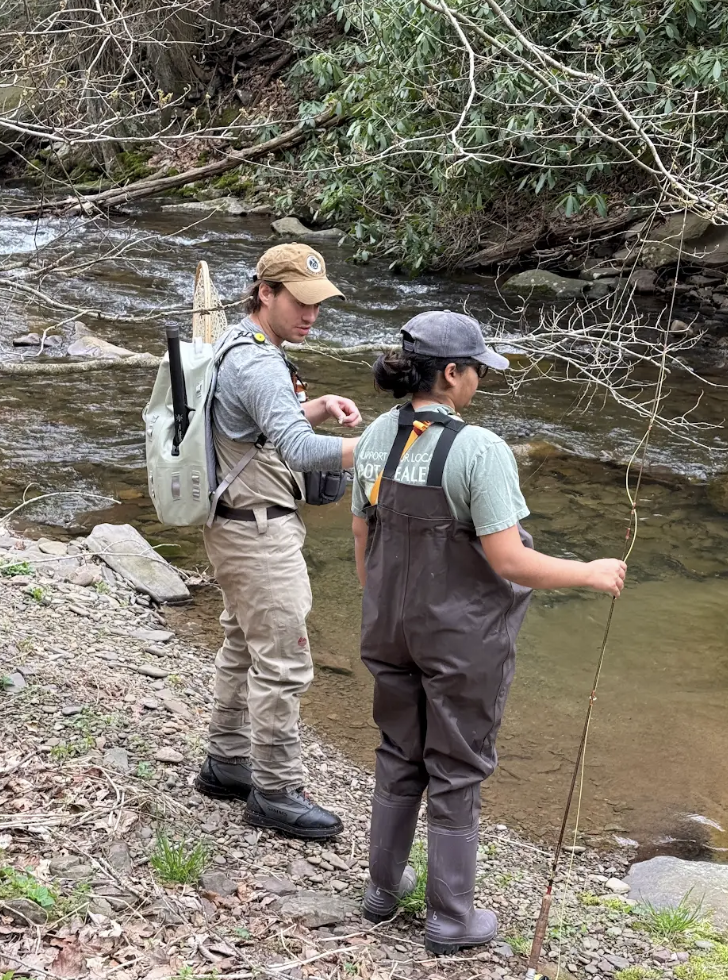 Two people in waders stand on a rocky riverbank with fishing gear, facing the water and preparing to fish in the stream.