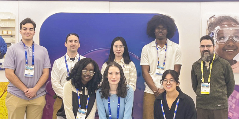 A group of eight people pose indoors in front of a colorful backdrop at what appears to be a conference or event, all wearing name badges.