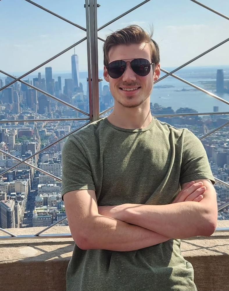 A man in sunglasses and a green shirt stands with arms crossed on an observation deck, with a city skyline and skyscrapers in the background.