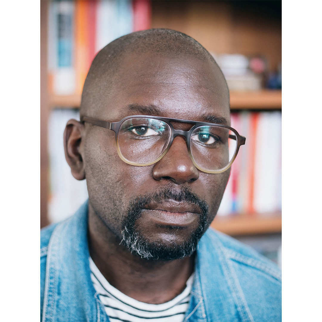 A man with glasses and a short beard wears a denim jacket and striped shirt, standing in front of a bookshelf.