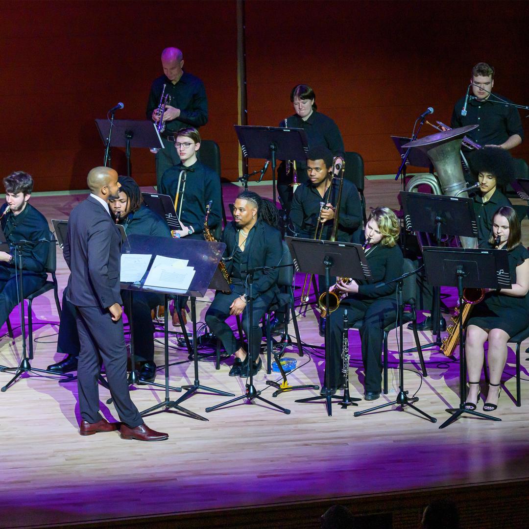 A conductor leads a seated ensemble of musicians playing various wind and brass instruments on a brightly lit stage.