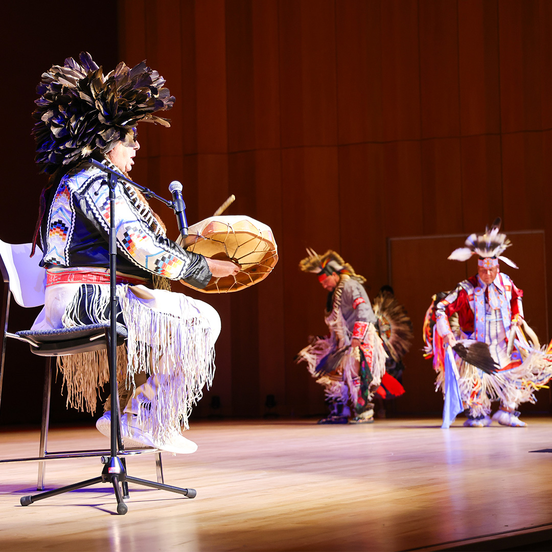 A seated performer in traditional regalia plays a drum and sings into a microphone while two dancers in feathered headdresses perform on a wooden stage.