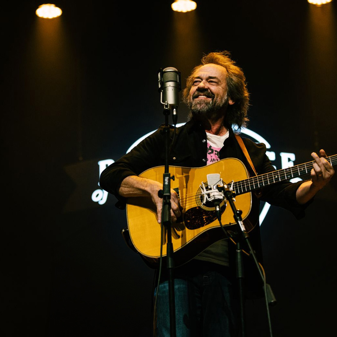 A man stands under stage lights, playing an acoustic guitar and singing into a microphone during a live performance.
