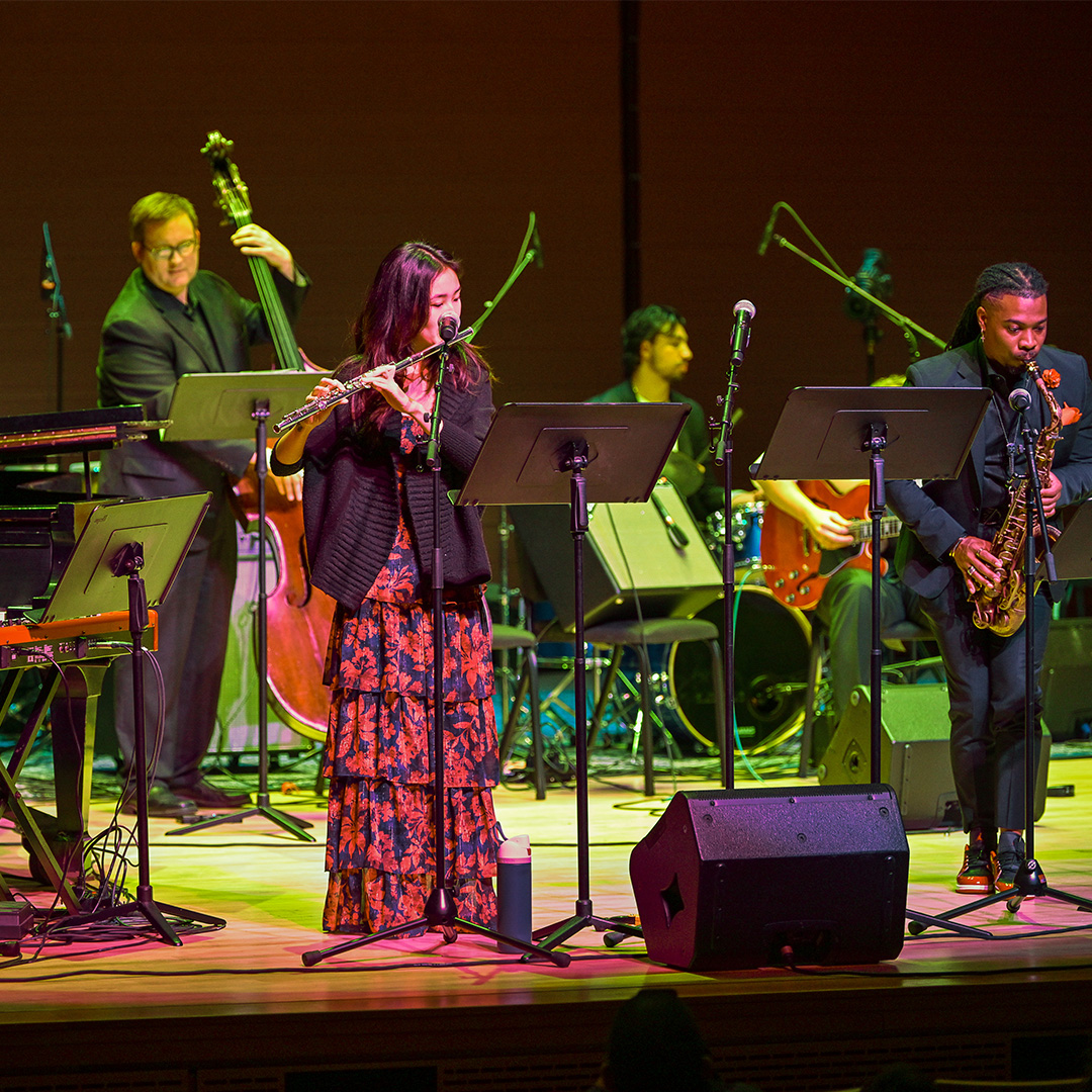 A jazz band performs on stage; musicians play the flute, saxophone, double bass, and drums, reading sheet music from stands.