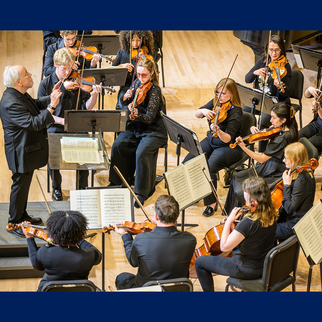 A conductor leads a group of violinists and string musicians during a performance, with sheet music stands visible on a wooden stage.