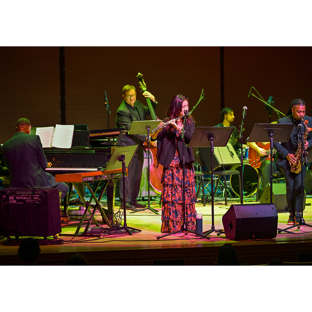 A jazz ensemble performs on stage with musicians playing piano, bass, keyboard, flute, saxophone, and drums under stage lighting.