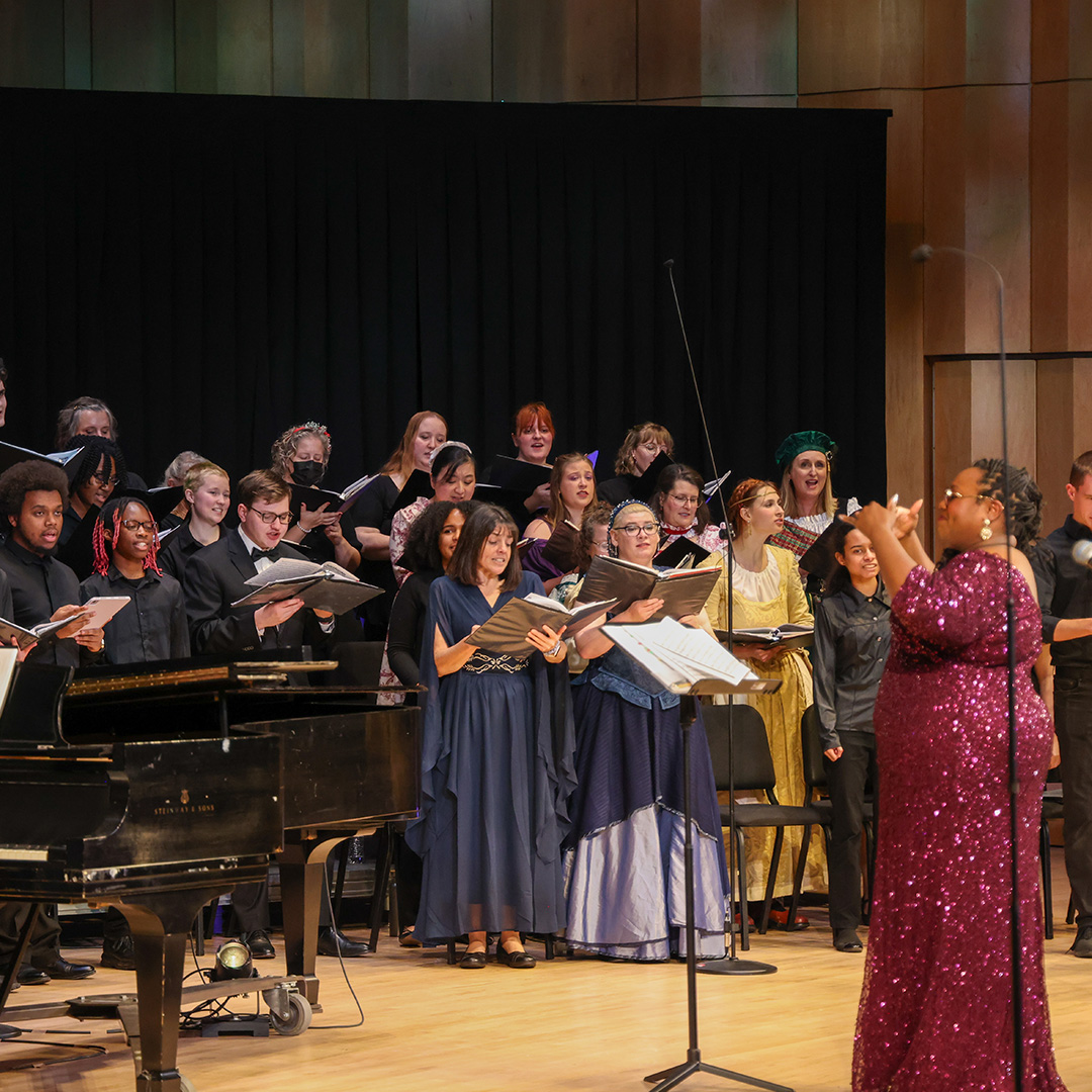 A choir in formal attire sings on stage with a conductor in a sequined dress leading them; a grand piano is visible on the left.