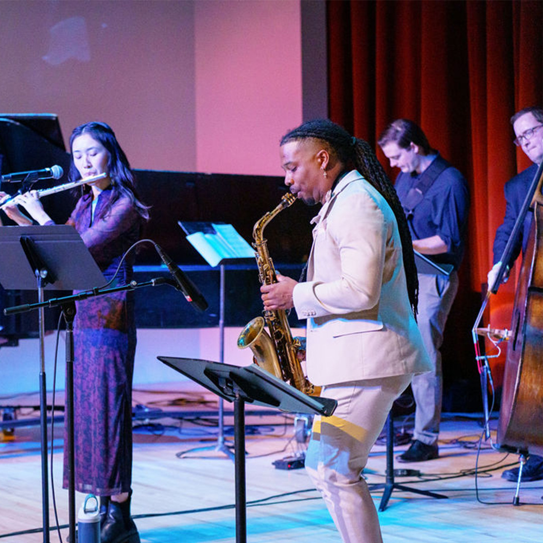 A group of four musicians perform on stage, playing the flute, saxophone, double bass, and keyboard, with music stands and red curtains in the background.