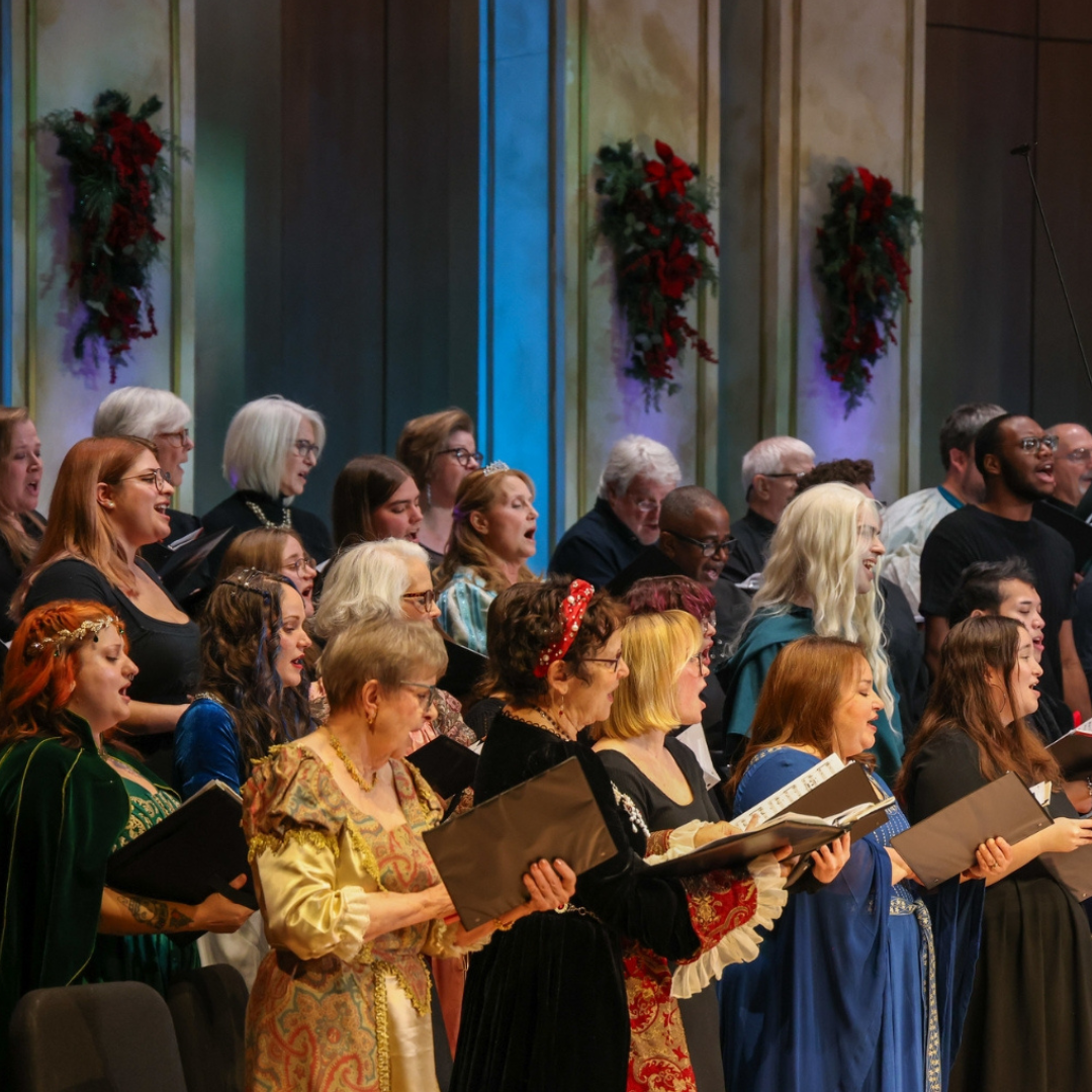 A group of people in colorful costumes sing in a choir on stage, with holiday wreaths hanging on the wall behind them.