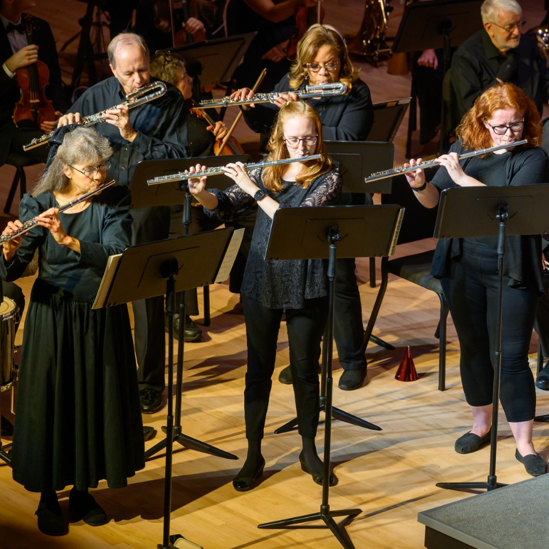 A group of musicians play flutes on a well-lit stage with music stands in front of them.
