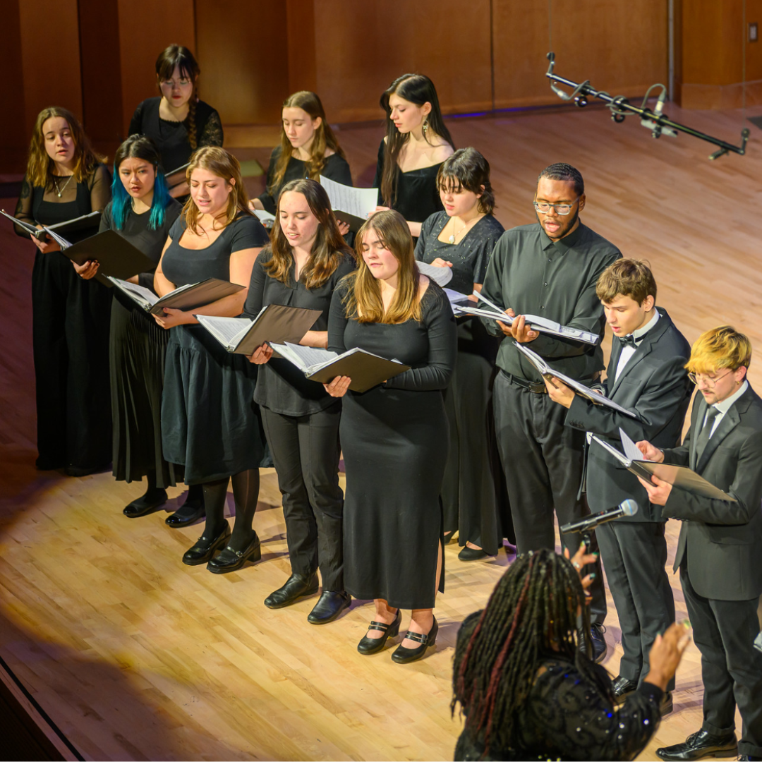 A choir of twelve people stands in a semi-circle on stage, holding sheet music and singing, with a conductor facing them and a microphone overhead.