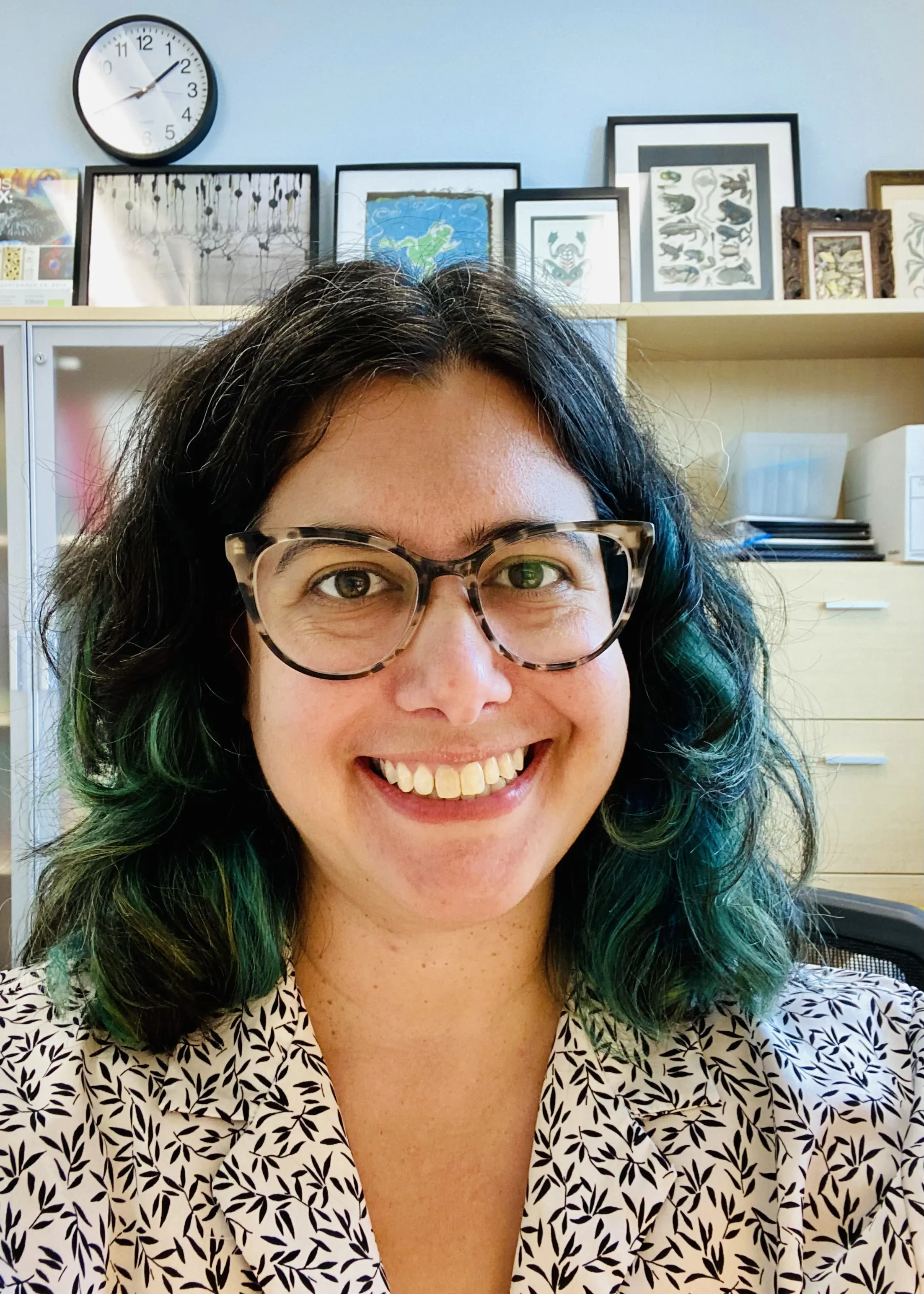 Me, Liz Leininger, looking at the camera and smiling. I have dark brown hair with parts dyed teal, wear tortise-shell pattern glasses and am wearing a white shirt with black leaf pattern