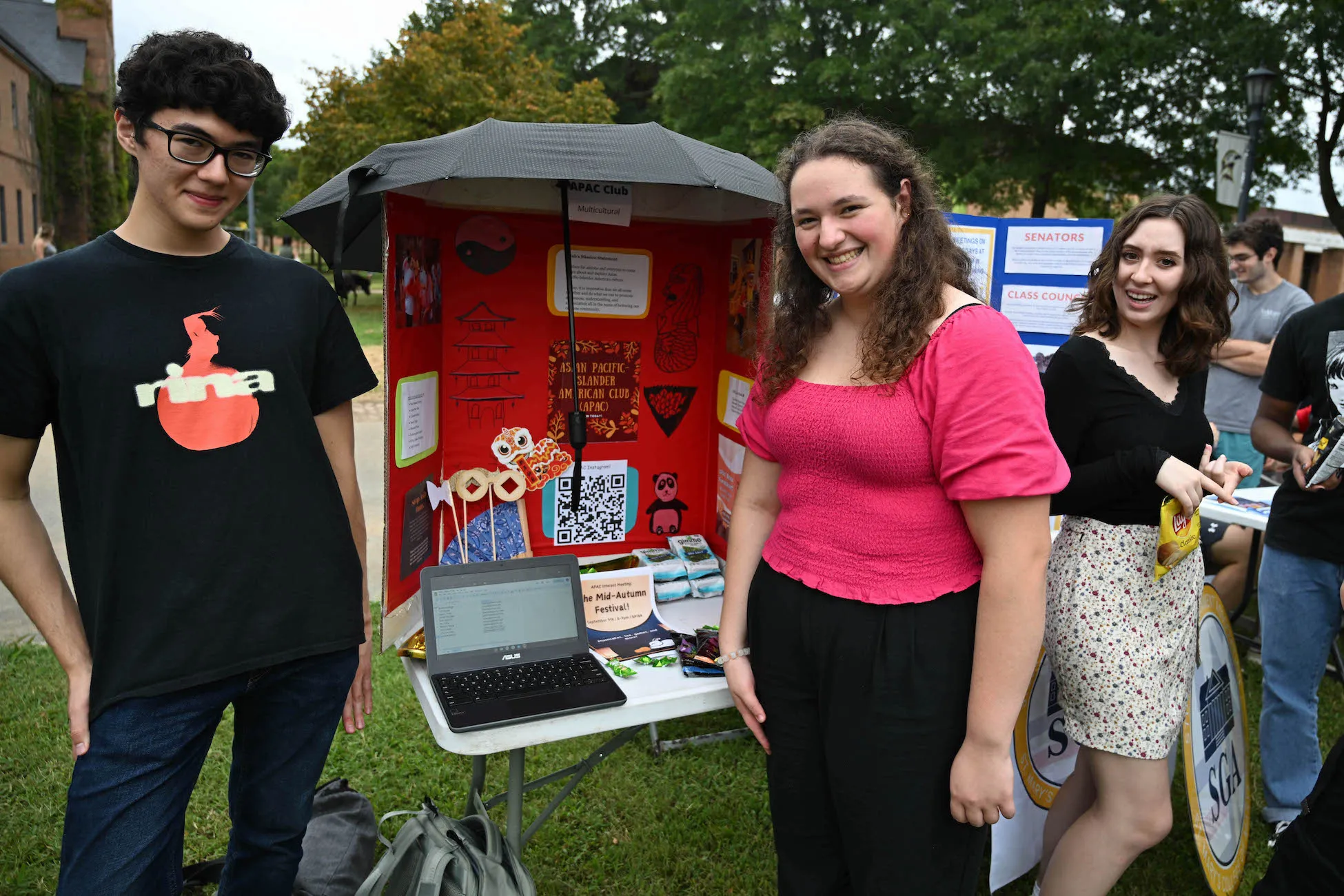 Three students posing in front of an Asian Pacific Islander American Culture (APAC) Club table
