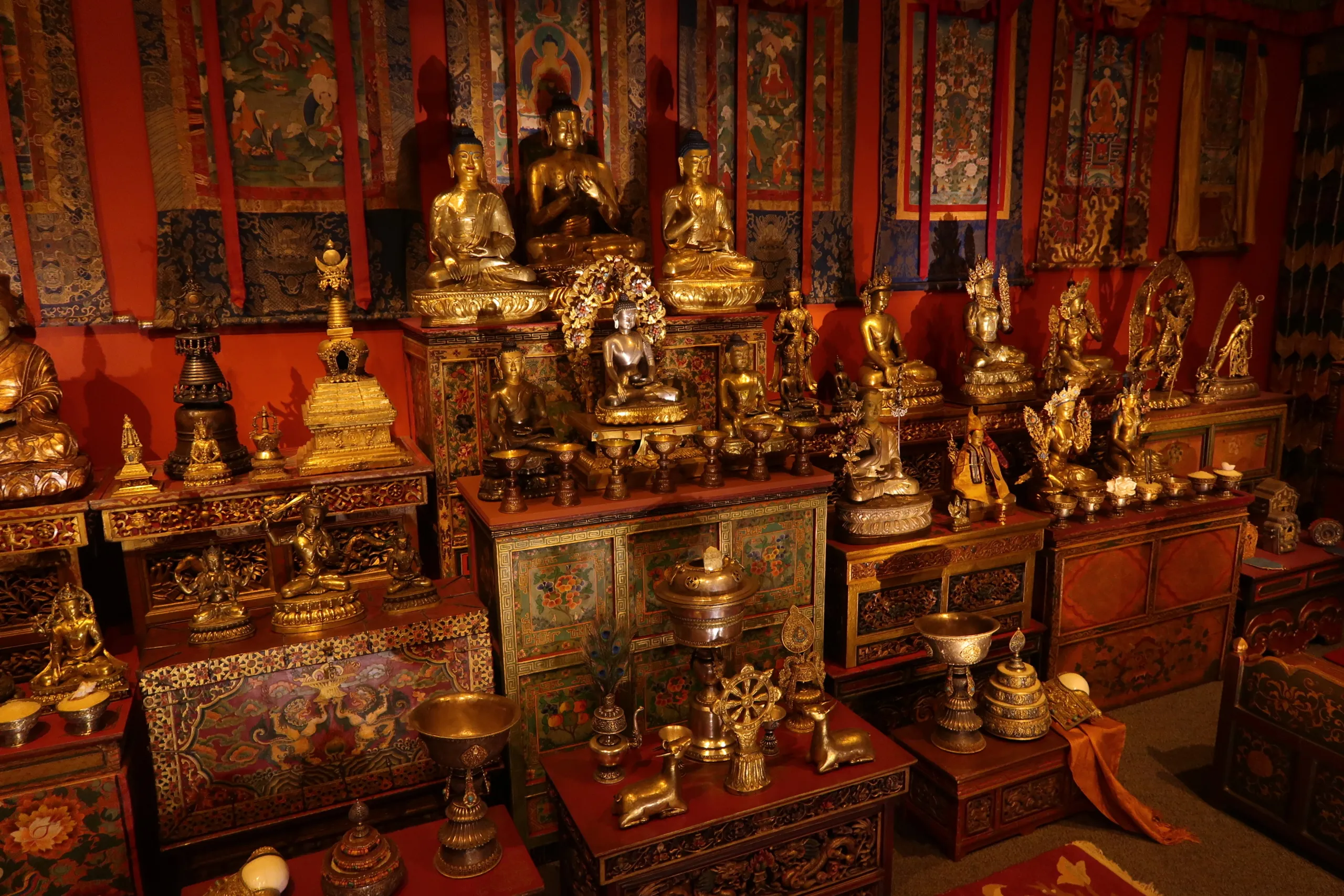 A richly decorated room filled with numerous golden statues and ornate artifacts, set against vibrantly painted walls with intricate details. The room conveys a sense of spiritual or cultural significance.