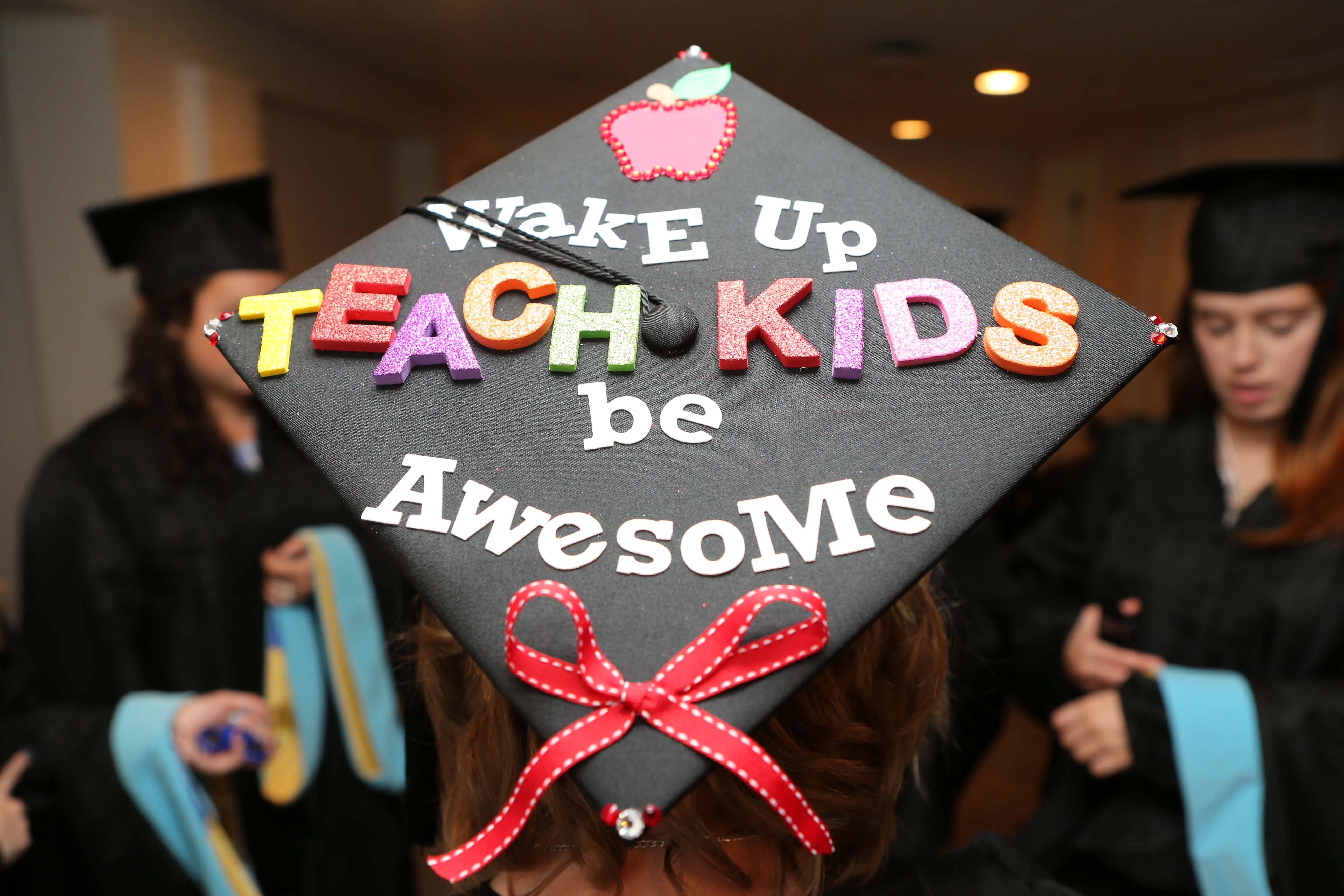 A graduation cap features colorful letters spelling "Wake Up Teach Kids be Awesome," adorned with an apple icon and a red ribbon. Students wearing black gowns are in the background.