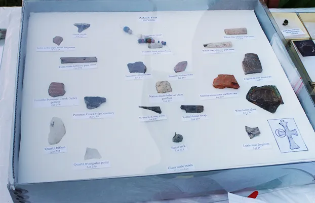 Collection of labeled archaeological artifacts displayed in a box. Items include stone fragments, pottery shards, a metal object, and glass, each with a brief description on white cards.