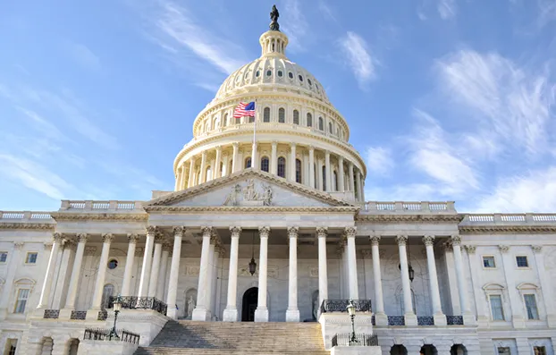 The image shows the United States Capitol building with its distinctive dome and a U.S. flag flying in front. The sky is partly cloudy, providing a backdrop to the neoclassical architecture of the historic structure.