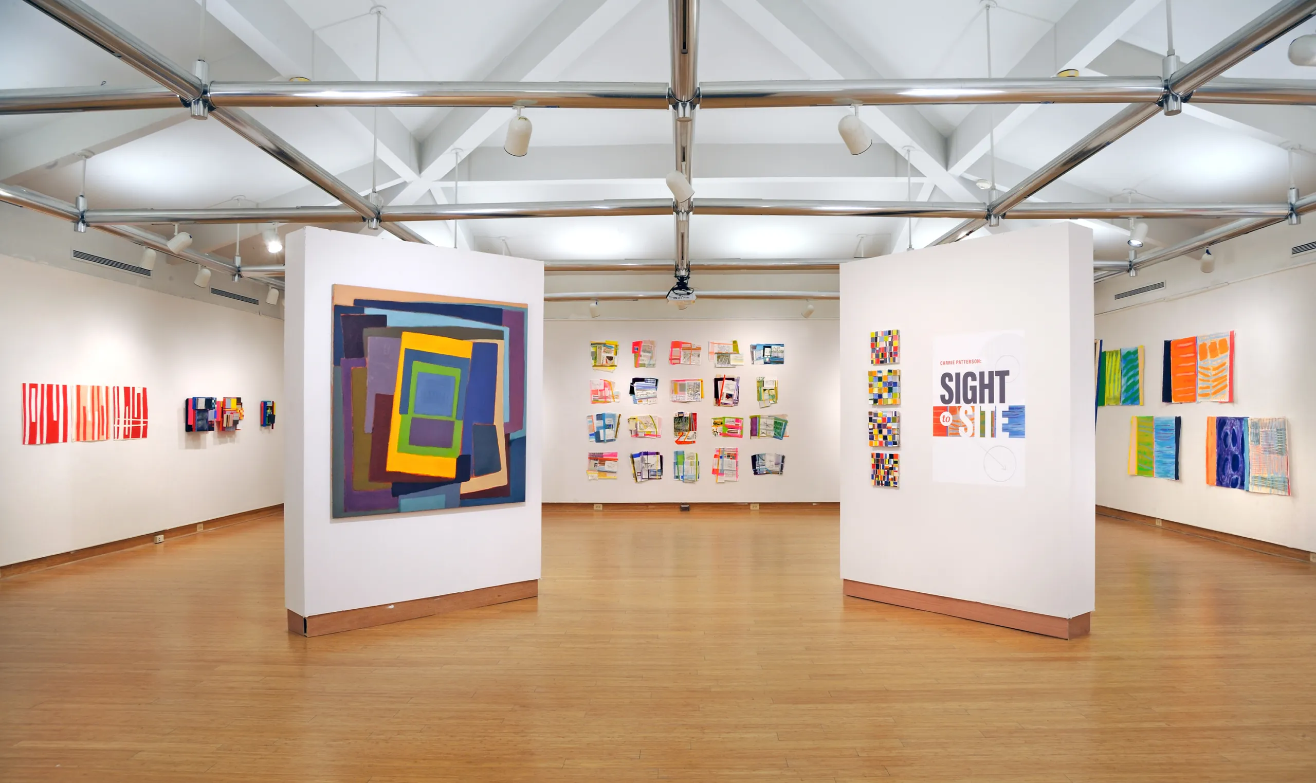 A contemporary art gallery features abstract artworks on display. Large colorful abstract paintings occupy central white walls, with smaller pieces arranged on surrounding walls. The space has a wooden floor and modern overhead lighting.