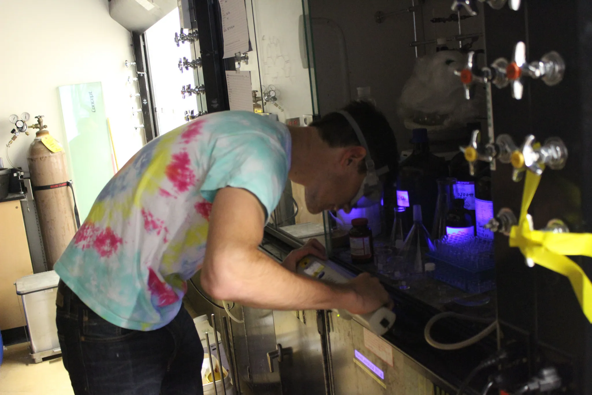 A person wearing safety goggles and a tie-dye shirt works in a laboratory. They are focused on using equipment inside a fume hood, surrounded by various instruments and bottles. Fluorescent lighting creates a blue glow in the workspace.