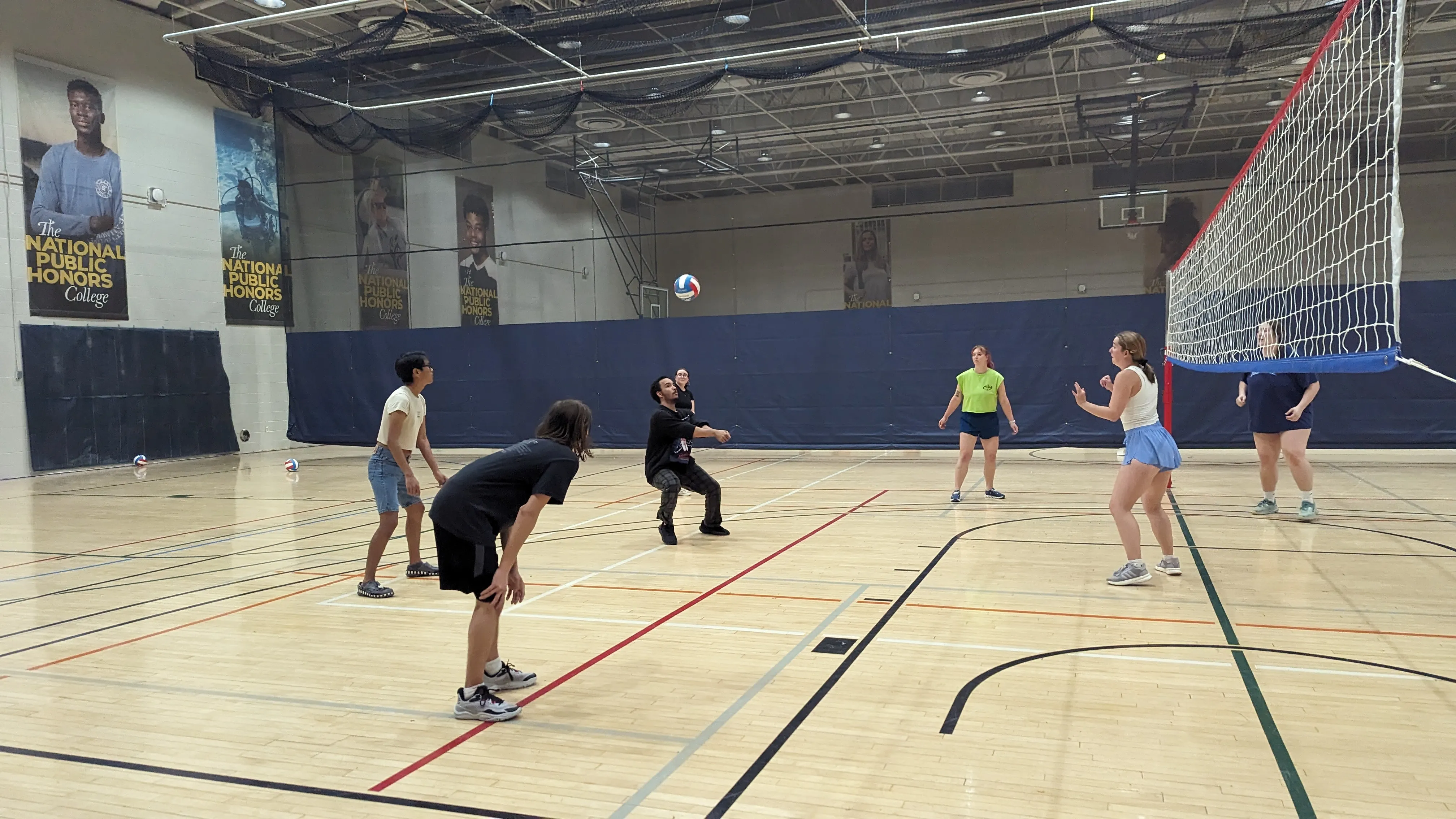 A group of six people play volleyball indoors on a gym court, with one player preparing to hit the ball over the net.