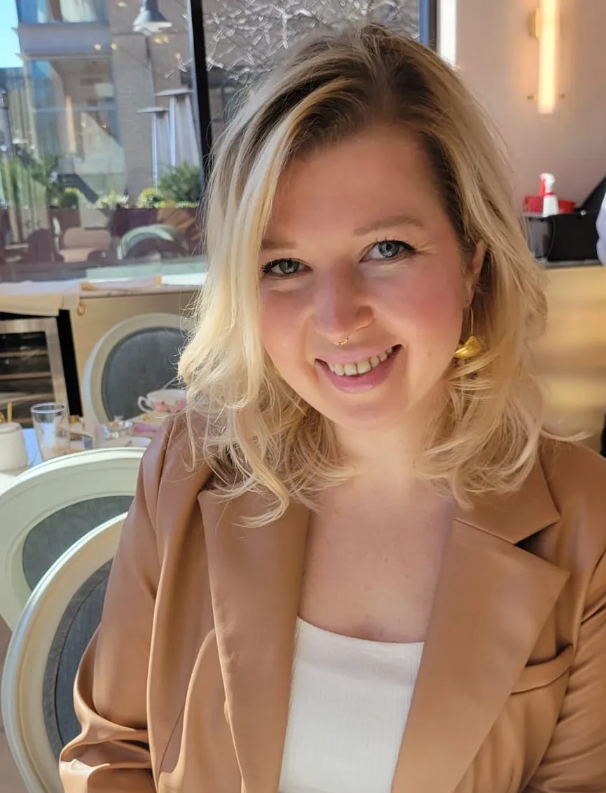A woman with blonde hair, wearing a tan blazer and white top, smiles while sitting at a table in a sunlit café or restaurant.