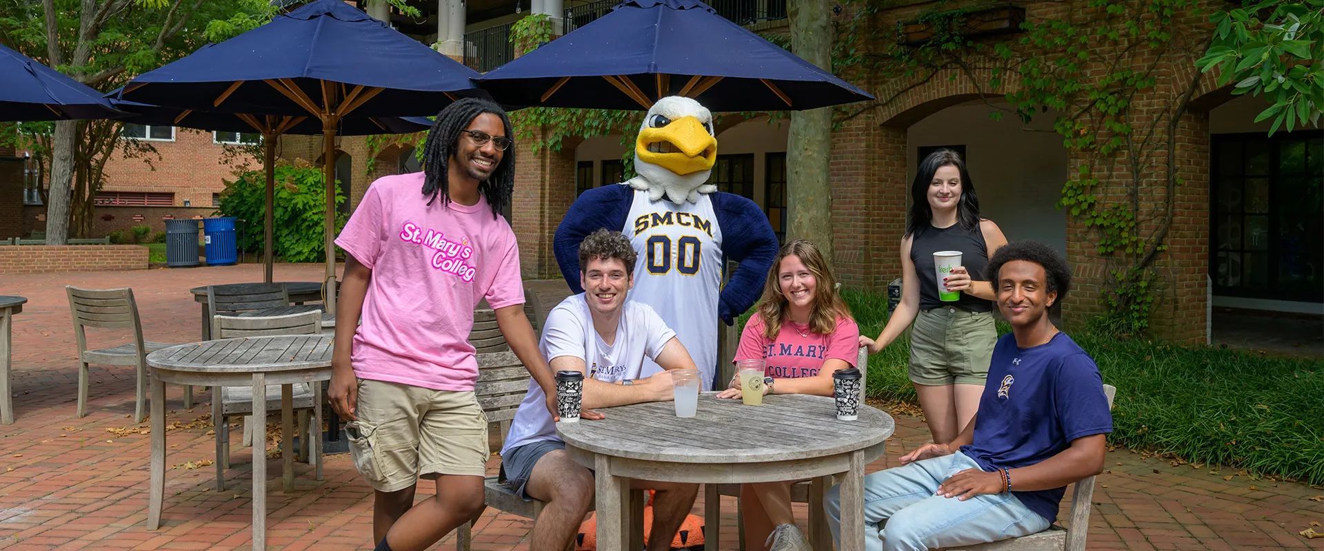 Five students and a person in a bald eagle mascot costume pose and smile at outdoor tables with drinks, under large umbrellas, on a brick patio near academic buildings.