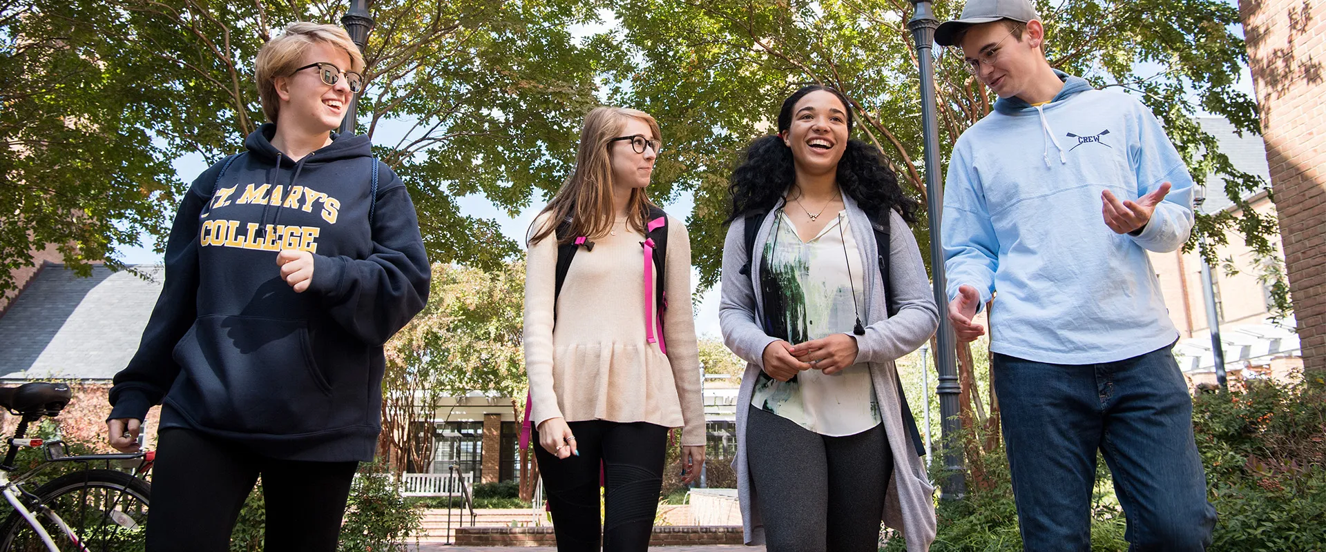 Four college students walk together outdoors on a sunny day, conversing and smiling, with trees and campus buildings in the background.