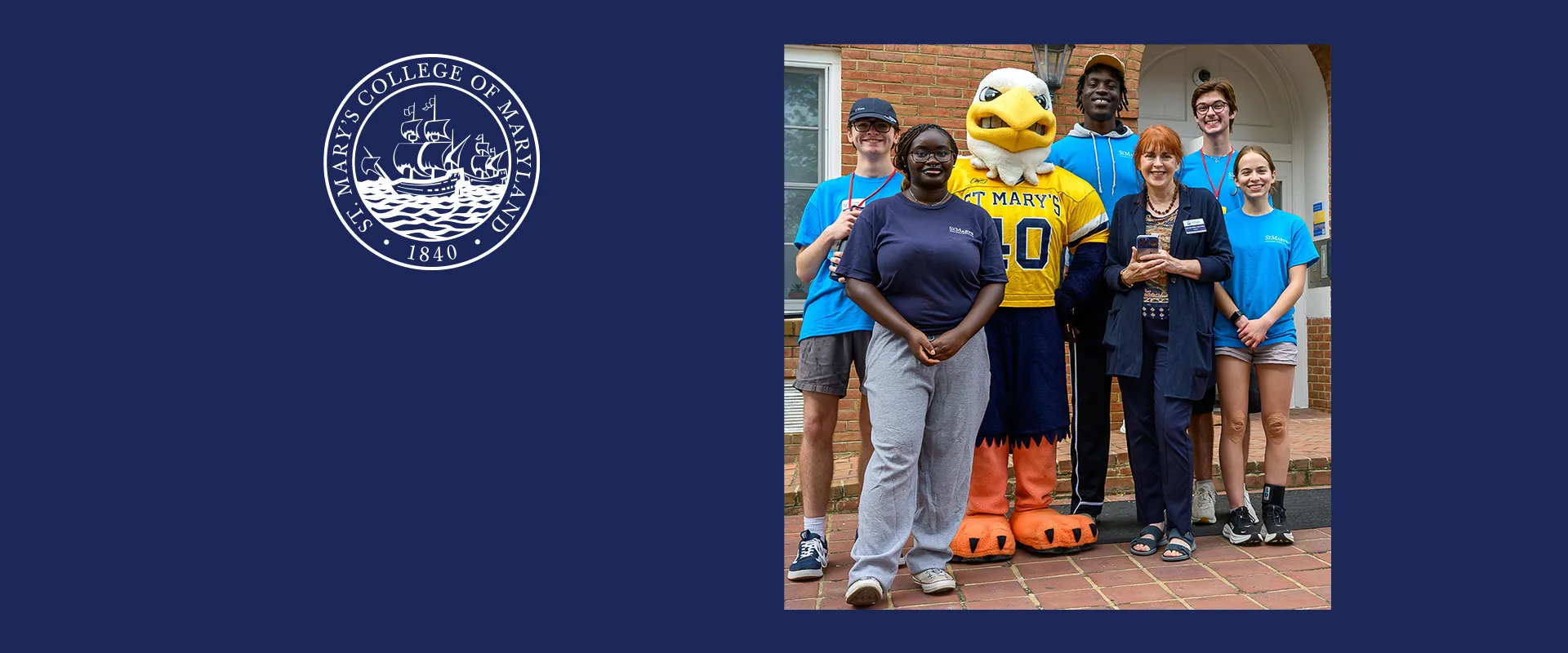 A group of six people, including a mascot in an eagle costume, pose and smile for a photo in front of a brick building at St. Mary’s College of Maryland.