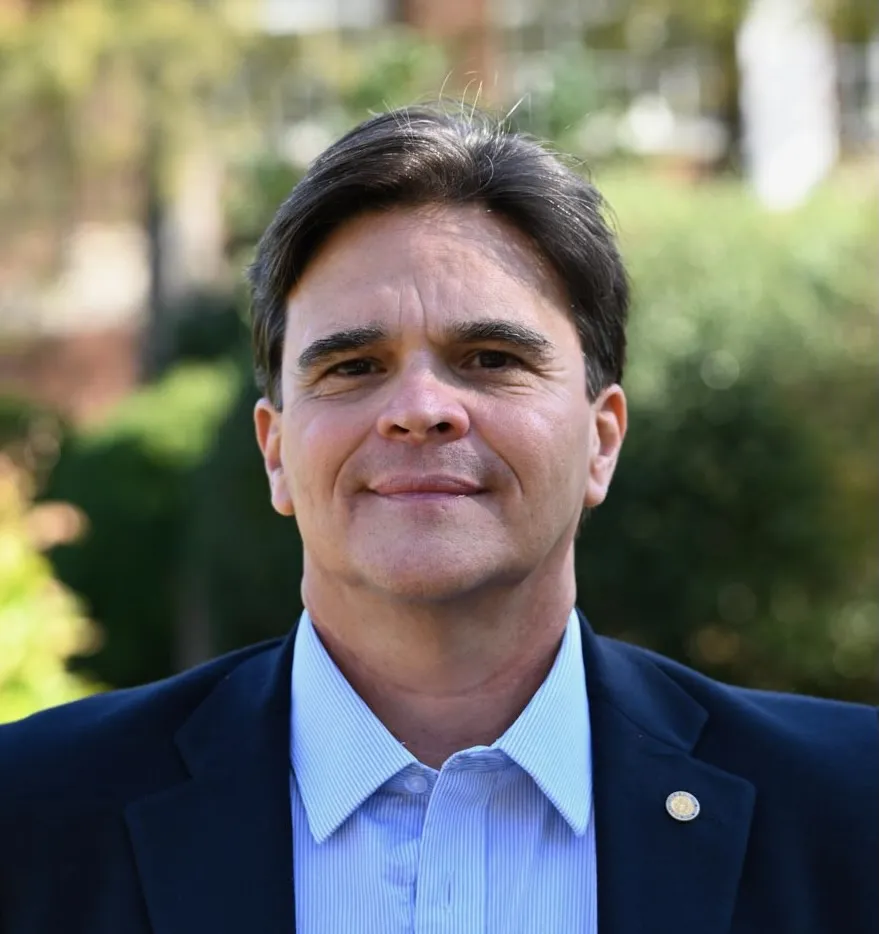 A man in a dark blazer and light blue shirt stands outside, facing the camera, with greenery and blurred buildings in the background.