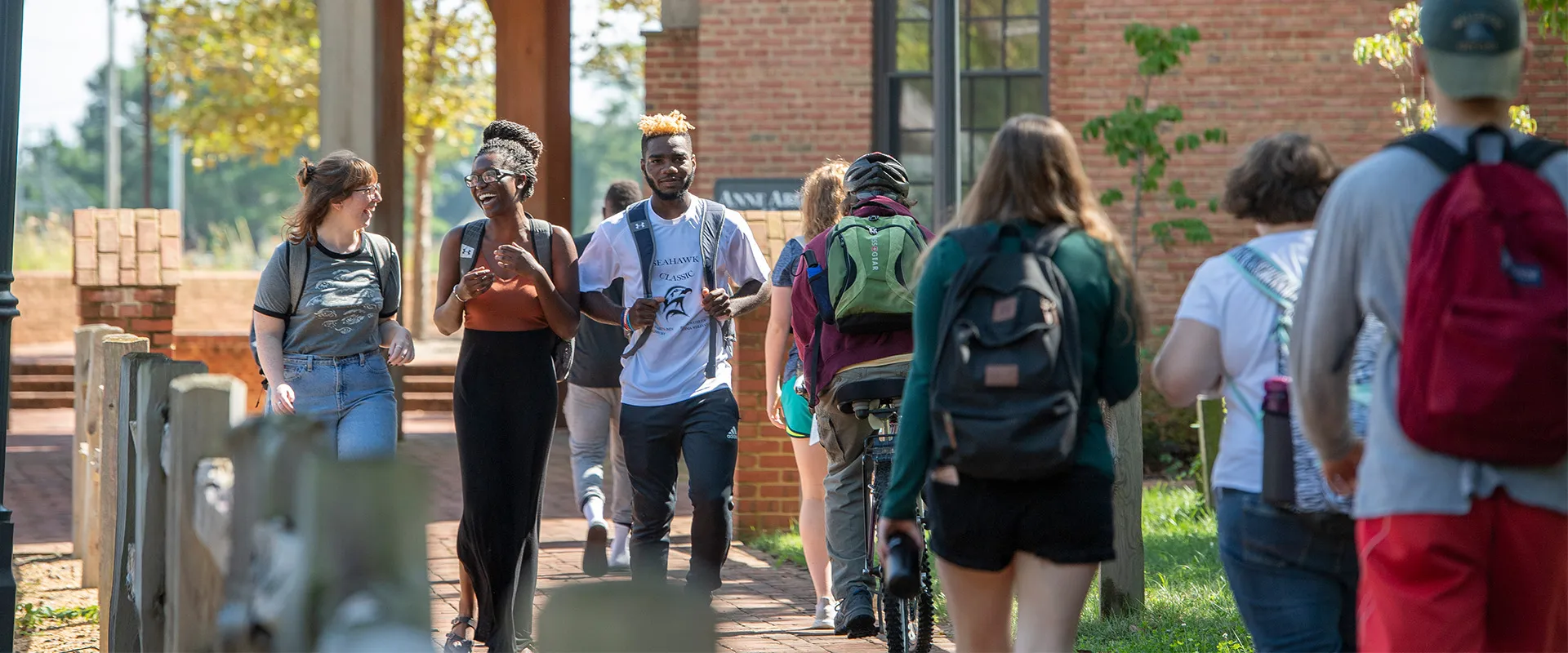 A group of students walk and talk outside on a sunny day near a brick building, some carrying backpacks and one person walking a bike.