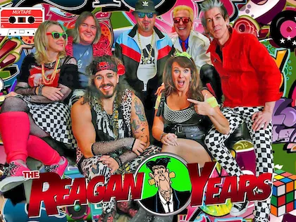 A group of eight adults in colorful, 1980s-themed costumes poses in front of a vibrant, graphic backdrop with "The Reagan Years" text at the bottom.