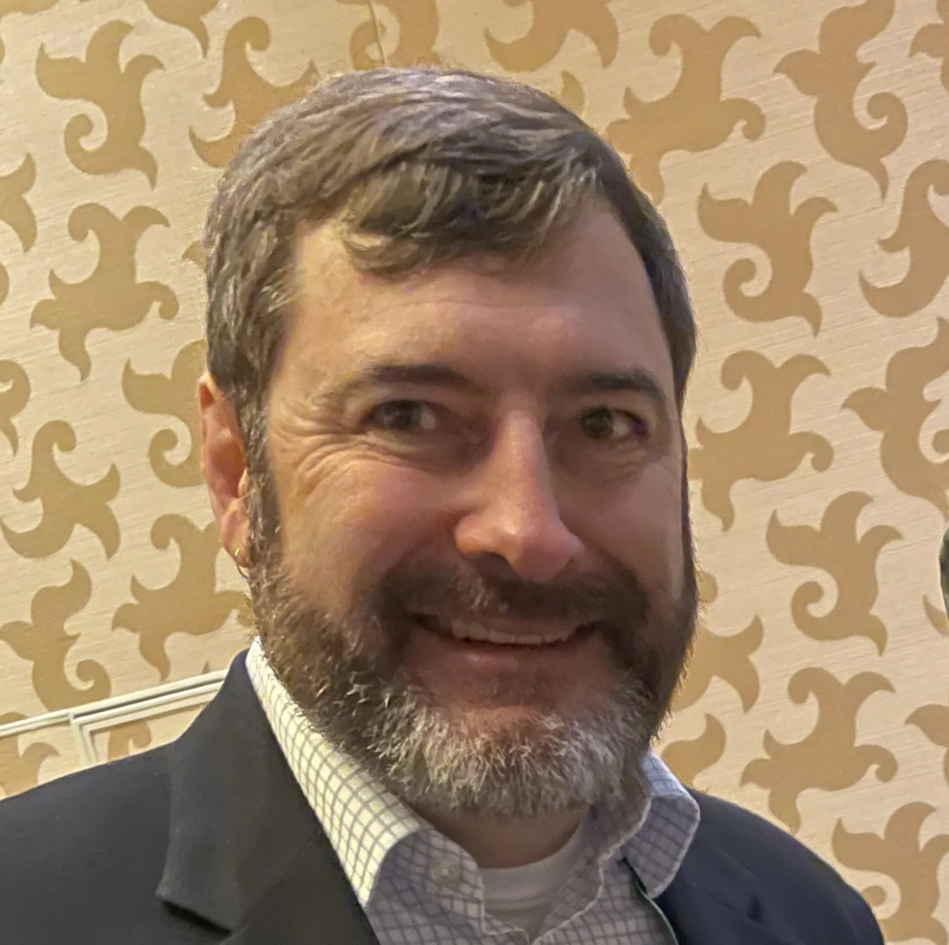 A man with brown hair and a beard, wearing a white shirt and black jacket, stands indoors in front of a beige patterned wallpaper.