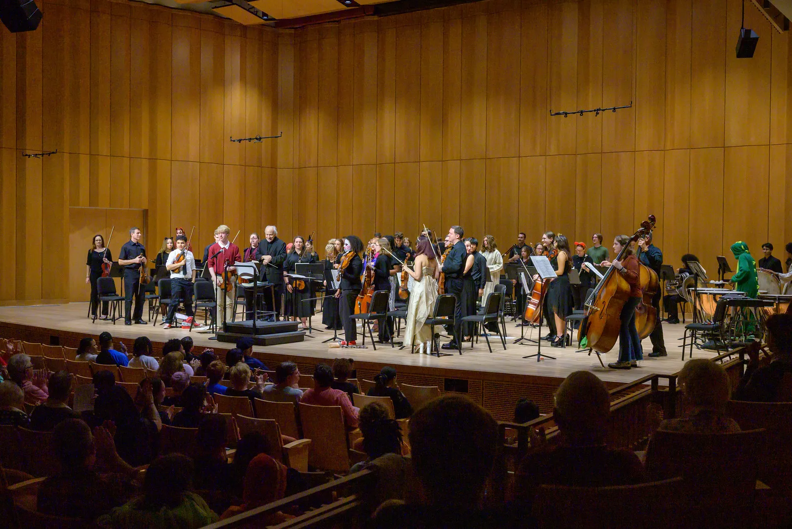 A symphony orchestra stands on stage with instruments, facing an applauding audience in a concert hall with wooden walls.