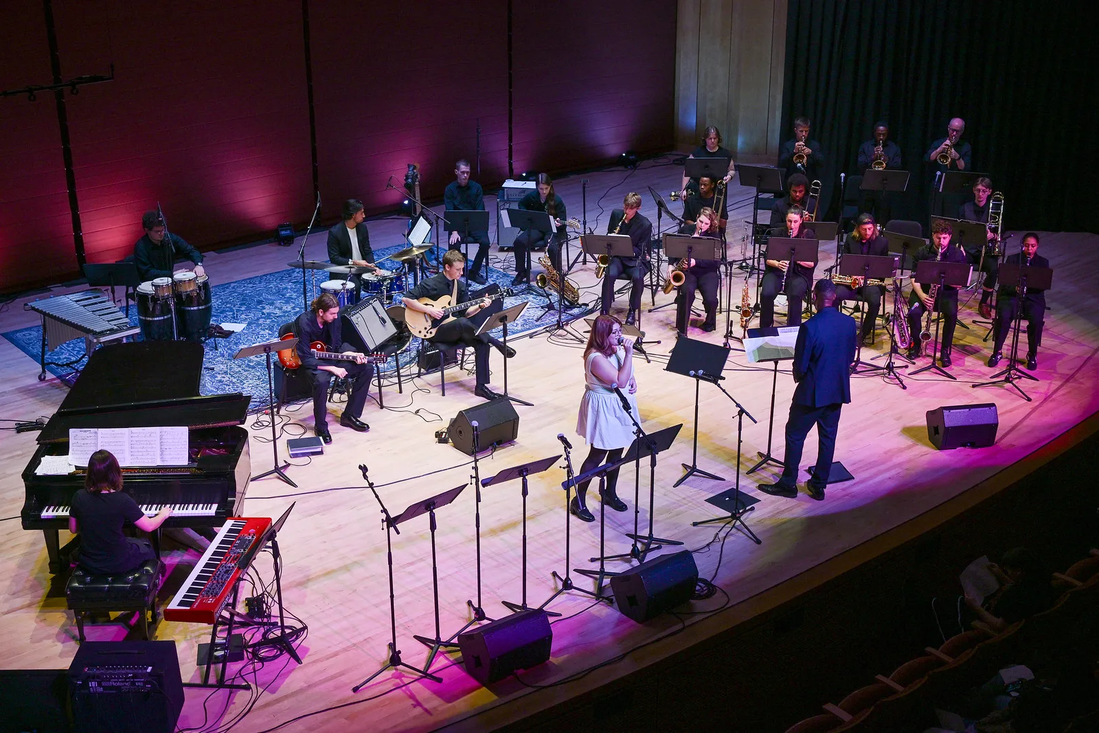 A large jazz band with multiple musicians performs on stage, featuring a singer and conductor in the front center, with various instruments including piano, drums, and brass.