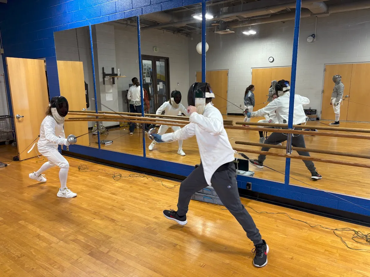 Two people in fencing gear spar in a gym with wooden floors and mirrored walls, while others observe in the background.